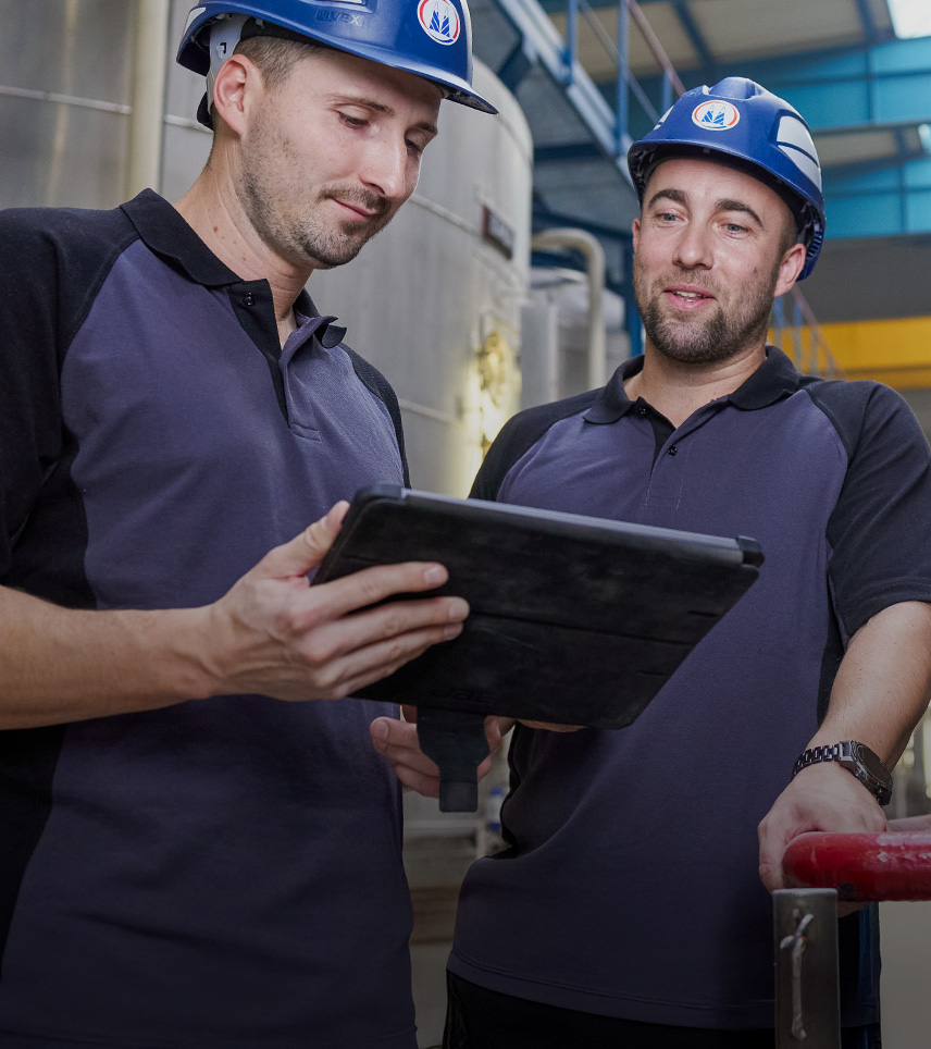 Zwei Mitarbeiter mit Schutzhelmen prüfen Daten auf einem Tablet in einer Industrieanlage.