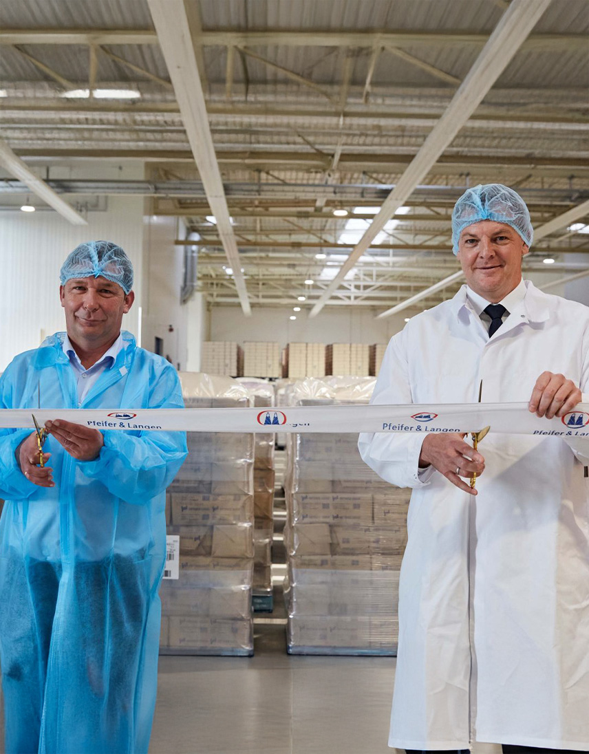 Zwei Personen in Schutzkleidung und Haarnetzen stehen in einer Lager- oder Produktionshalle und halten ein Band mit Firmenlogo. Beide haben eine Schere in der Hand, im Hintergrund sind Paletten mit verpackten Produkten zu sehen.