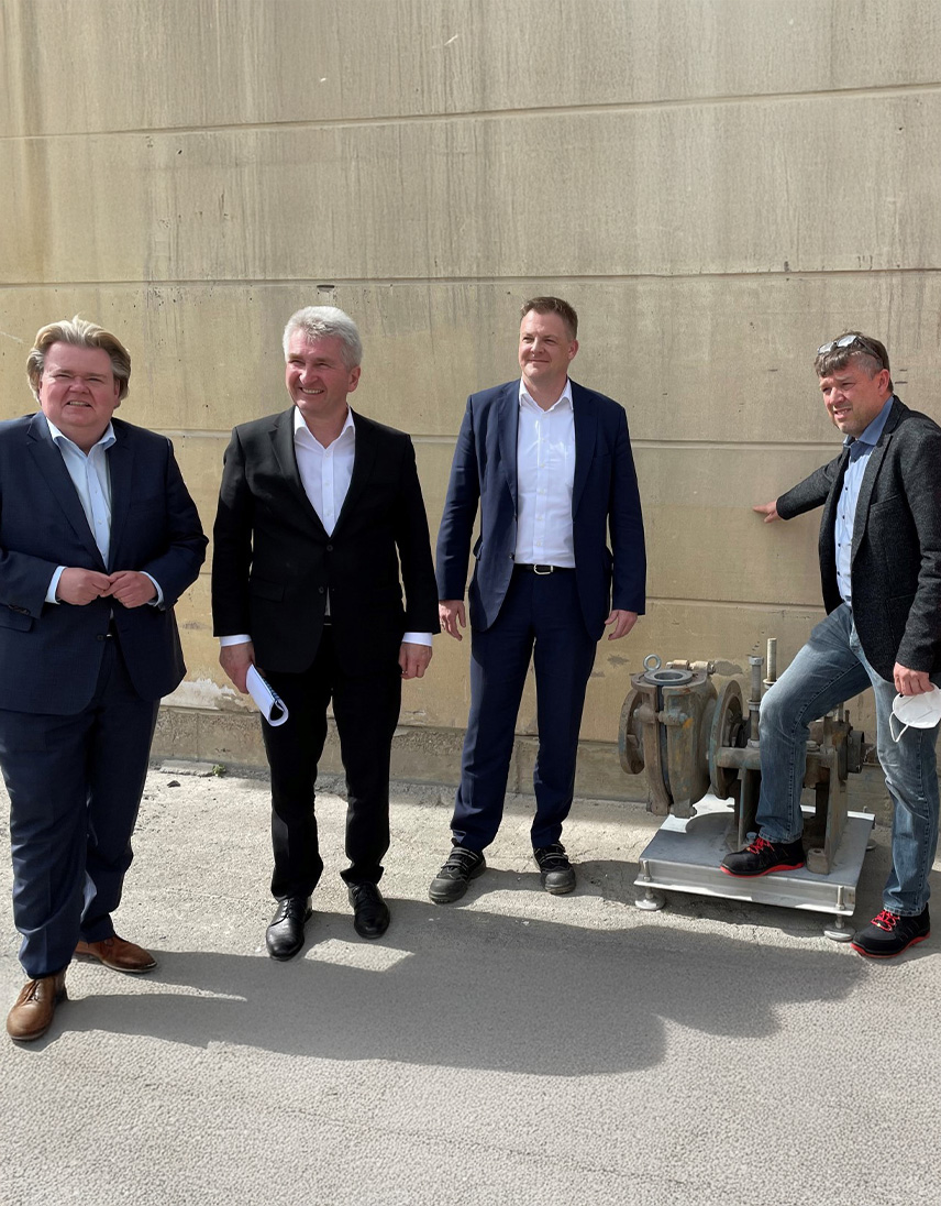 Vier Personen stehen vor einer Betonwand. Eine Person zeigt auf eine Anlage oder technische Vorrichtung am Boden, während die anderen danebenstehen.