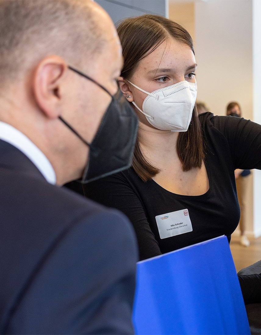 Eine junge Frau mit weißer Schutzmaske und Namensschild hält eine blaue Mappe unter dem Arm und zeigt auf etwas außerhalb des Bildes. Im Vordergrund ist ein Mann mit schwarzer Maske und Anzug zu sehen.