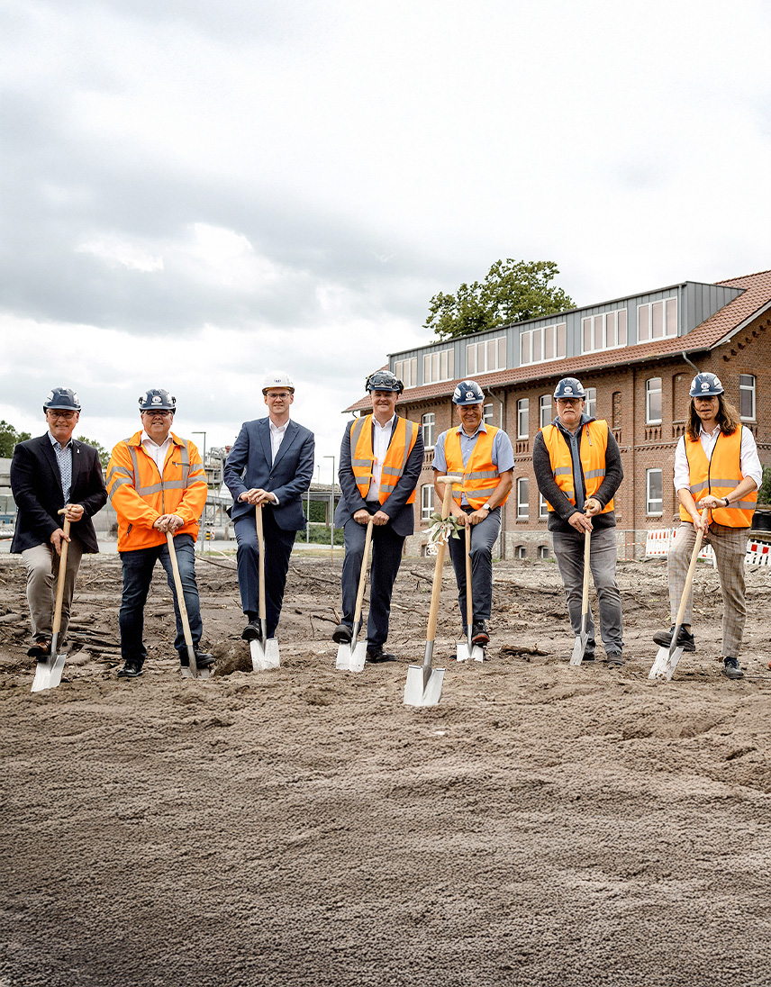 Acht Personen mit Schutzhelmen und teilweise Warnwesten stehen mit Spaten auf einer Baustelle.