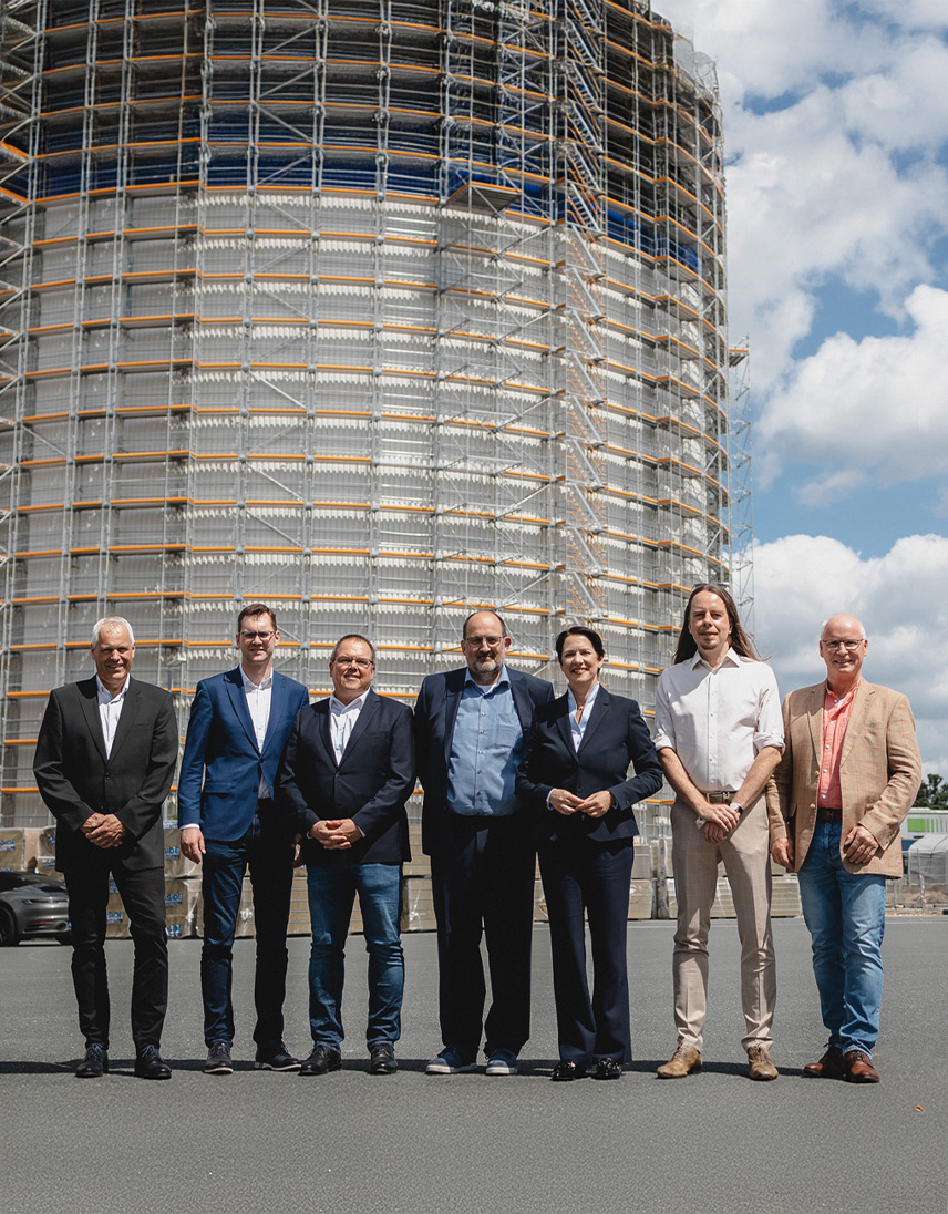 Sieben Personen stehen vor einem großen Industriesilo mit Gerüst auf einem Werksgelände.