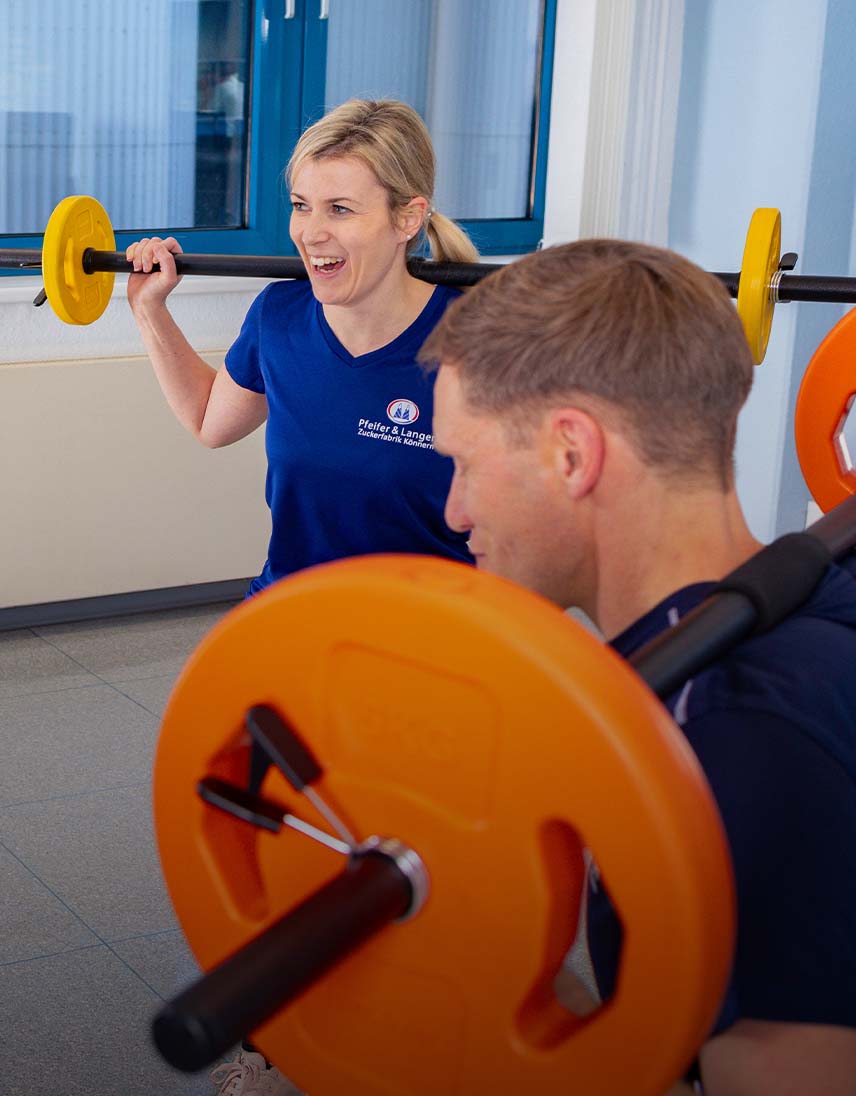 Zwei Personen machen gemeinsam Krafttraining mit Langhanteln in einem Fitnessraum.