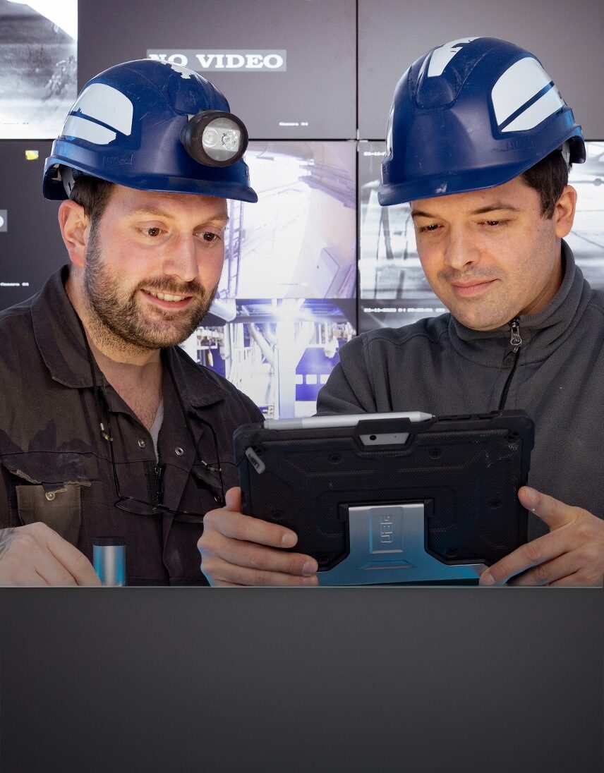 Zwei Männer mit Schutzhelmen schauen gemeinsam auf ein Tablet in einer industriellen Umgebung.