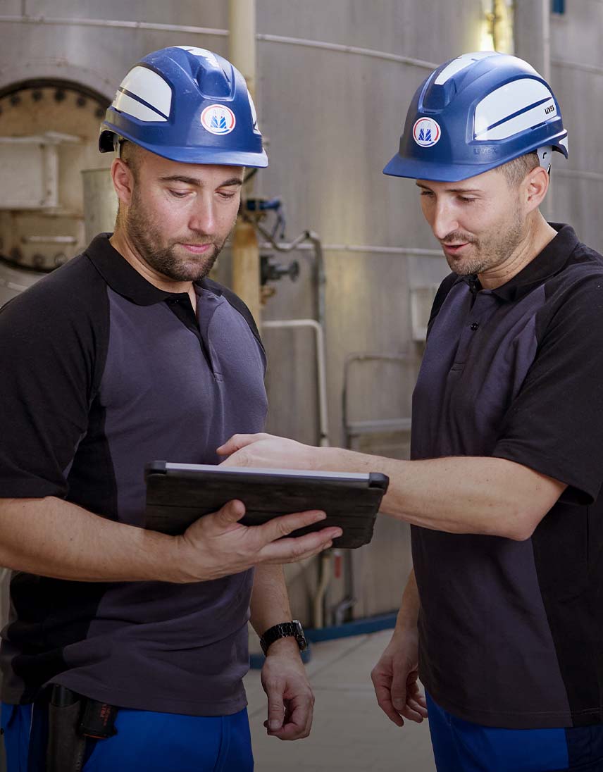 Two employees wearing safety helmets look at a tablet in an industrial facility.
