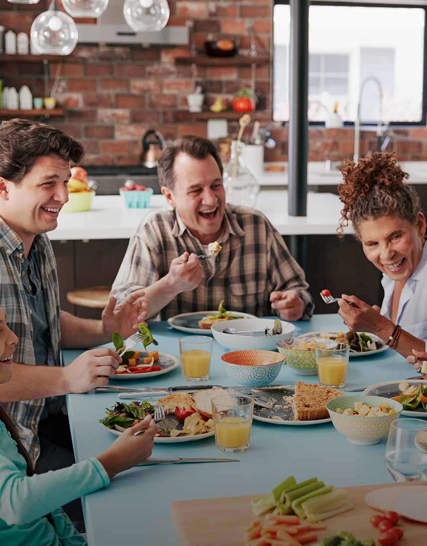 Vier Personen, darunter ein Kind, sitzen gemeinsam an einem Tisch in einer modernen Küche, essen und lachen, während verschiedene Gerichte und Getränke auf dem Tisch stehen.