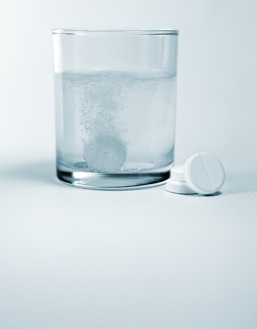 Brausetablette löst sich in einem Glas Wasser auf, daneben liegen weitere Tabletten.