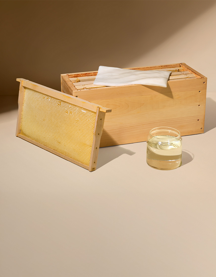 Holzbeute mit Wabenrahmen und Glas mit heller Flüssigkeit auf einer hellen Oberfläche.