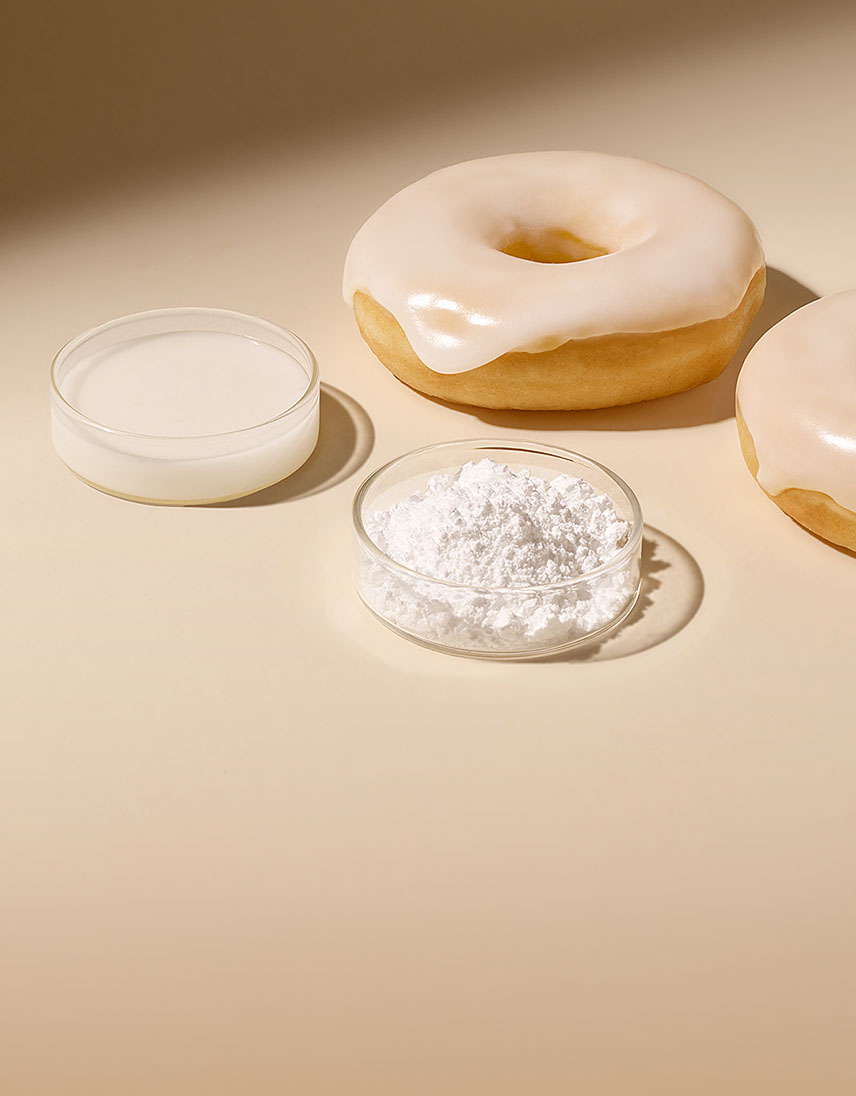 Zwei Donuts mit weißer Zuckerglasur liegen neben kleinen Schalen mit Puderzucker und flüssiger Zuckerglasur auf hellem Hintergrund.