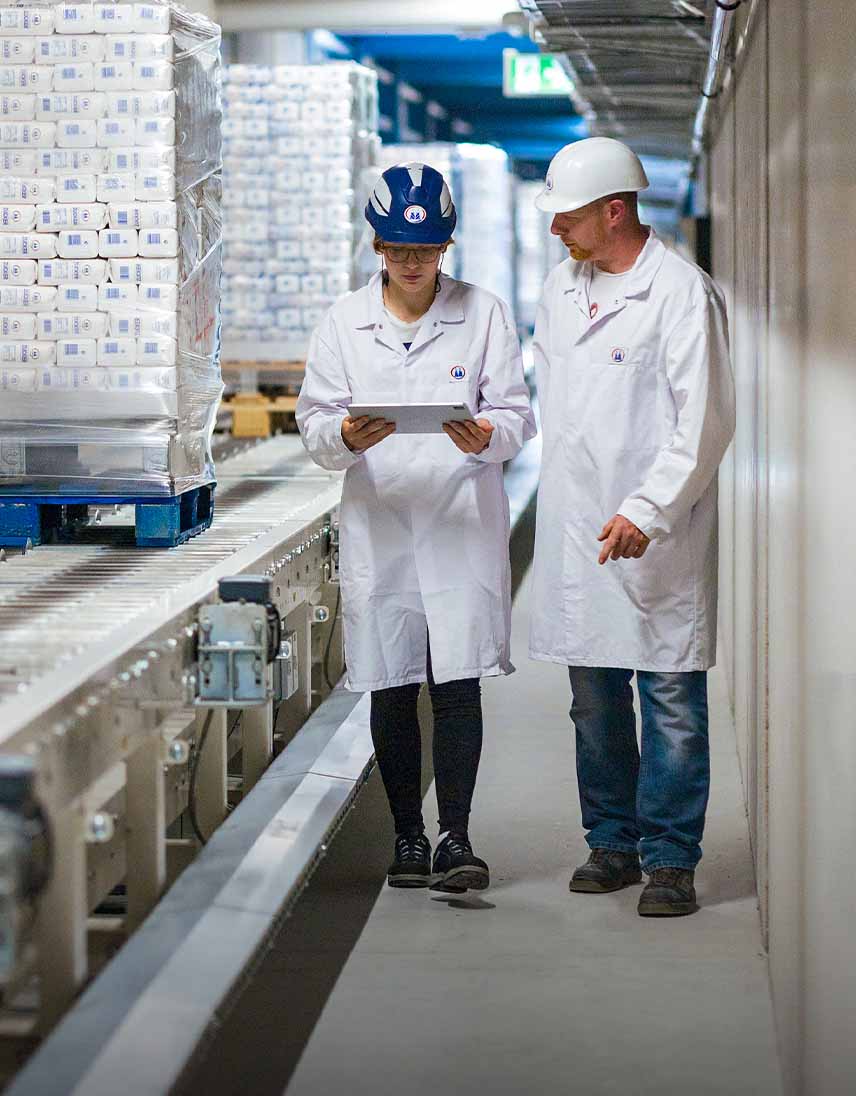 Zwei Personen in Schutzkleidung gehen durch eine Produktionshalle und schauen gemeinsam auf ein Tablet neben einer Förderanlage.