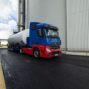 Blauroter Tanklastwagen fährt zwischen großen Industriesilos auf einem Werksgelände.