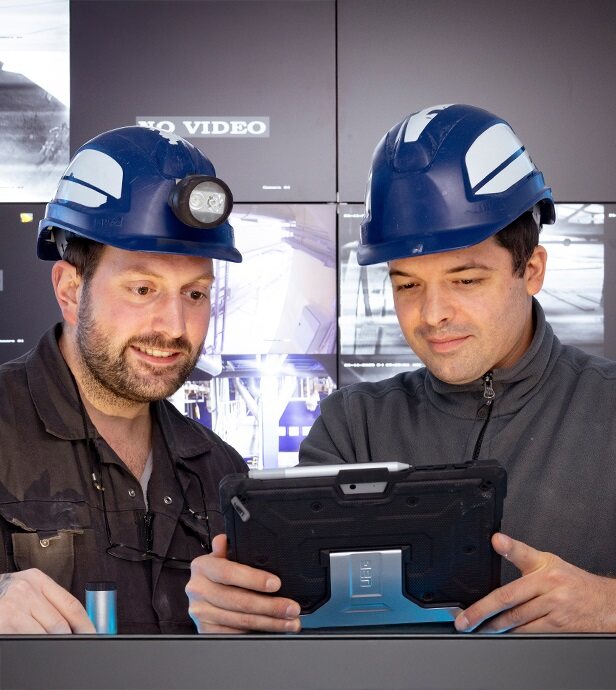 Zwei Männer mit Pfeifer und Langen Schutzhelmen schauen gemeinsam auf ein Tablet in einer industriellen Umgebung.