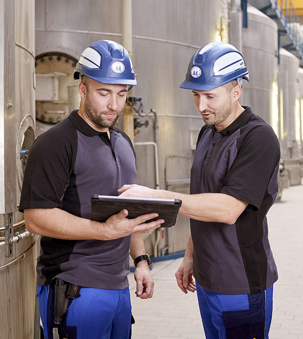 Zwei Mitarbeiter mit Schutzhelmen prüfen Daten auf einem Tablet in einer Industrieanlage.