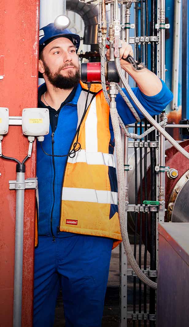 Arbeiter in Schutzkleidung überprüft Anlagen in einer Industriehalle zwischen Rohrleitungen und Maschinen.