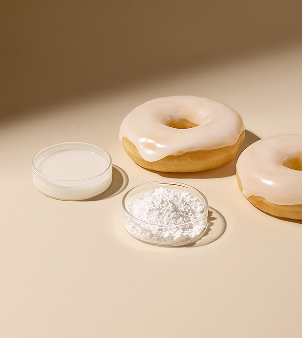 Zwei Donuts mit weißer Zuckerglasur liegen neben kleinen Schalen mit Puderzucker und flüssiger Zuckerglasur auf hellem Hintergrund.