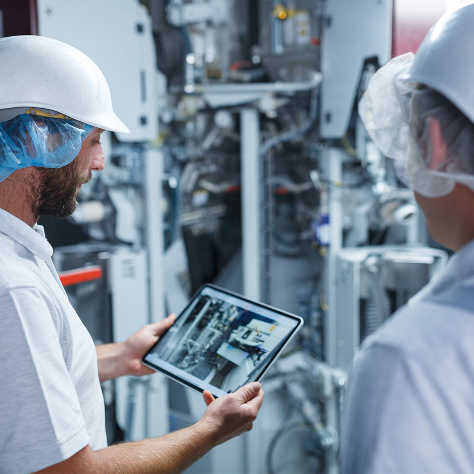 Zwei Mitarbeitende in Schutzkleidung betrachten ein Tablet vor einer industriellen Produktionsanlage.