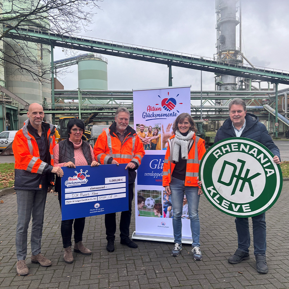 Fünf Personen stehen vor einer Industrieanlage; gemeinsam präsentieren sie einen Spendenscheck, während eine Person ein rundes Vereinsschild von ‚Rhenania Kleve‘ hält.