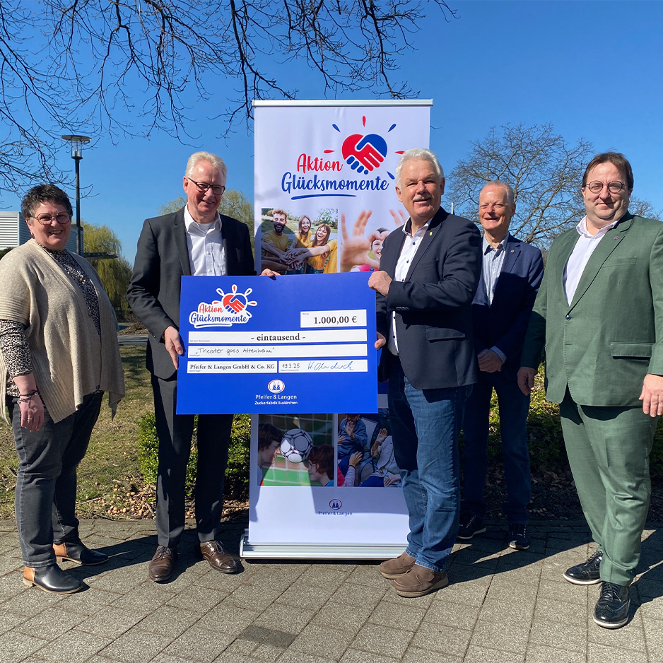 Fünf Personen präsentieren einen Spendenscheck vor einem Banner der Aktion Glücksmomente im Freien.