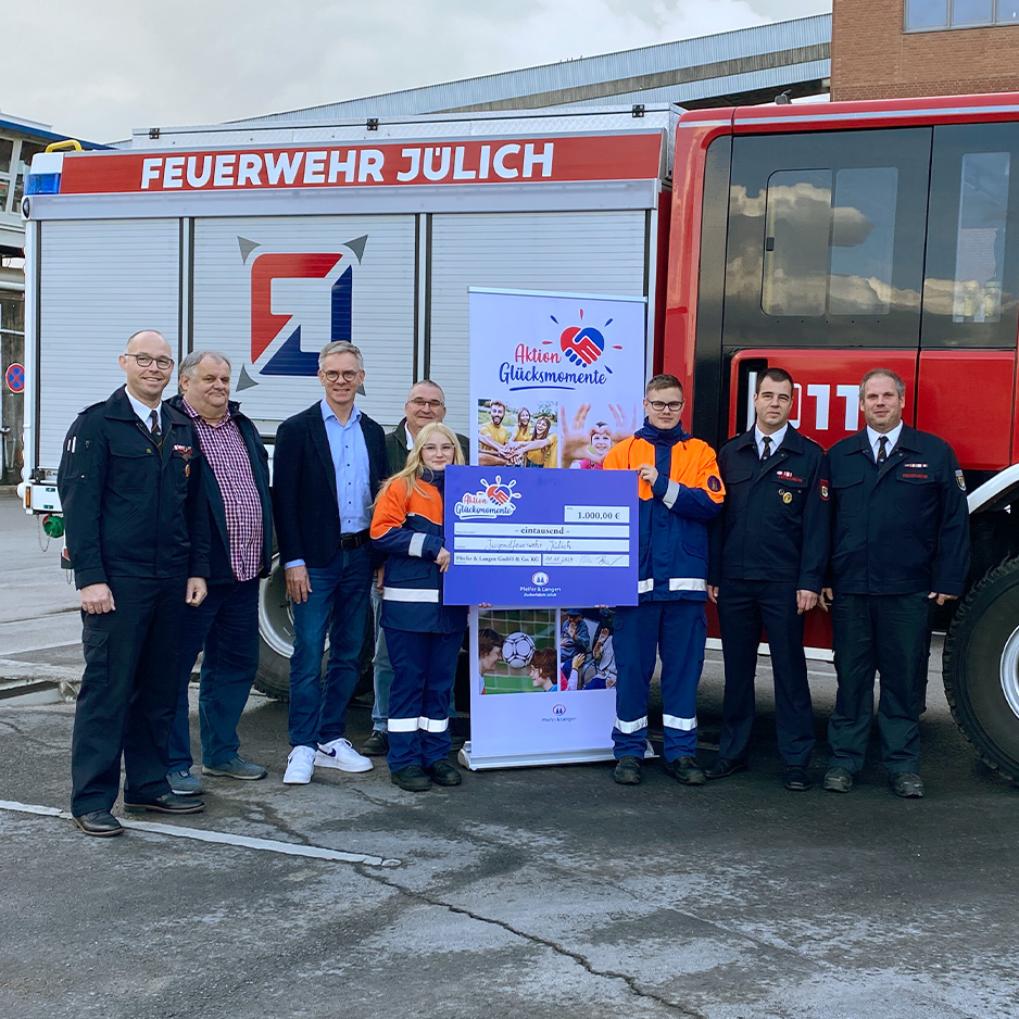 Gruppe präsentiert einen Spendenscheck vor einem Feuerwehrfahrzeug der Feuerwehr Jülich.
