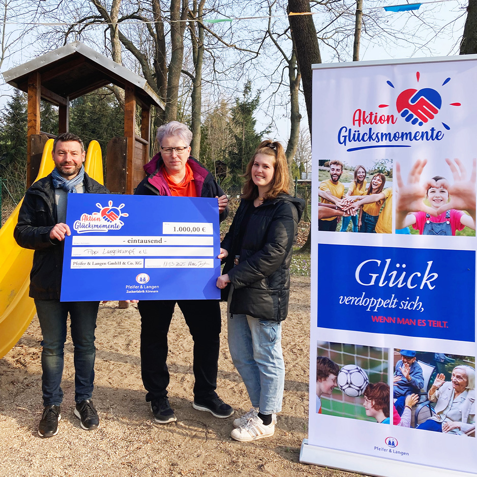 Drei Personen präsentieren einen Spendenscheck neben einem Banner der Aktion Glücksmomente auf einem Spielplatz.