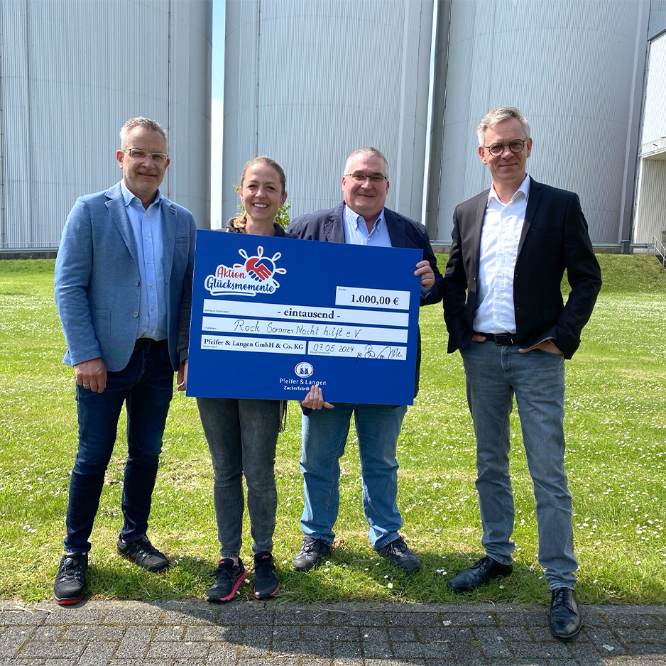 Vier Personen präsentieren einen Spendenscheck vor einem Banner der Aktion Glücksmomente im Freien.